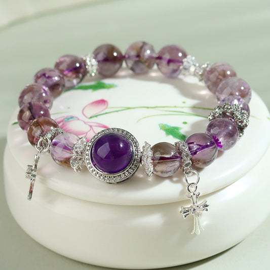 Amethyst crystal bracelet with cross charms resting on a white ceramic dish with floral painting — soft lavender light, serene and elegant